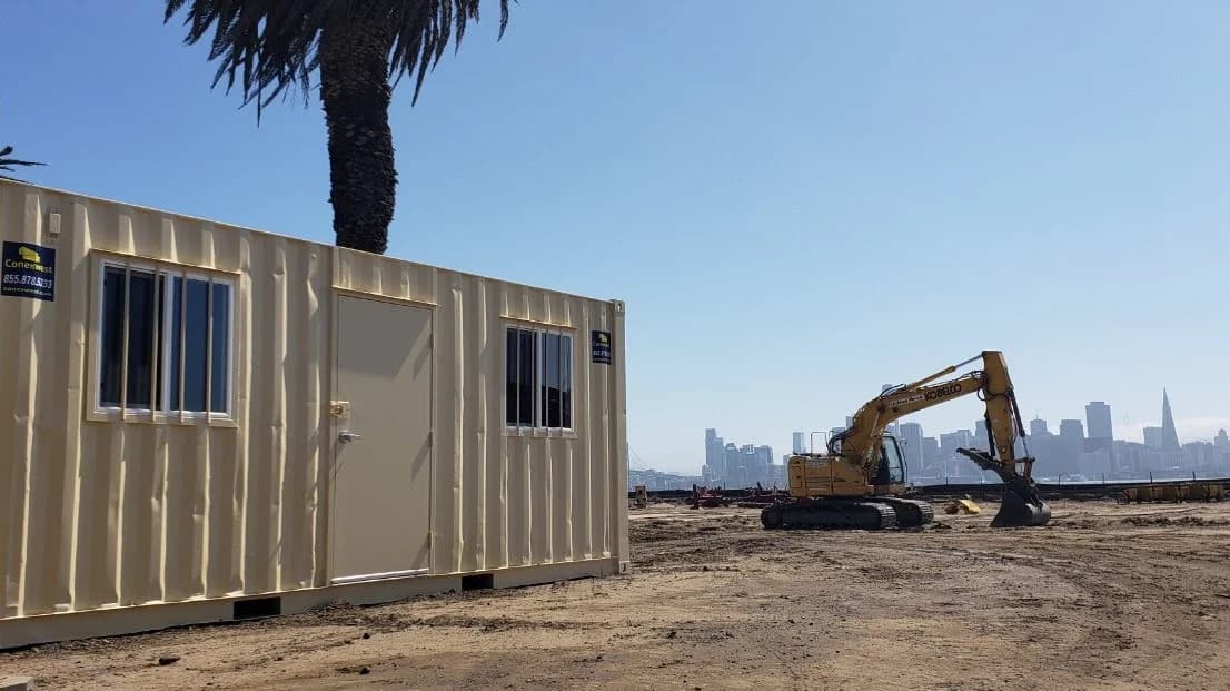 A Conexwest container with a personnel door and windows near an excavator parked on a San Francisco construction site