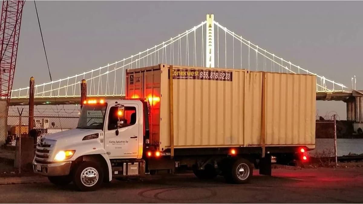 A Conexwest delivery truck transports a yellow shipping container
