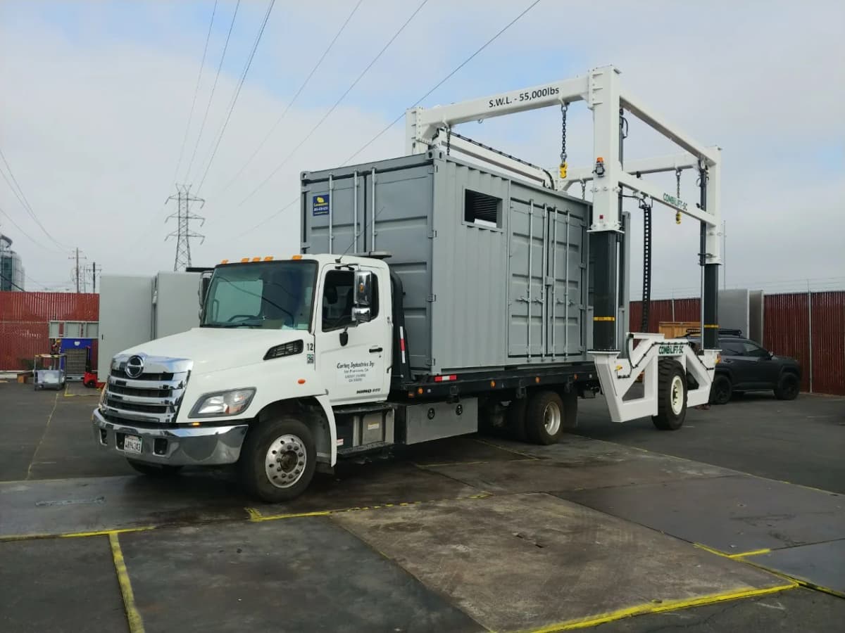 A white truck is transporting a large gray Conexwest container secured by a mobile lifting system.
