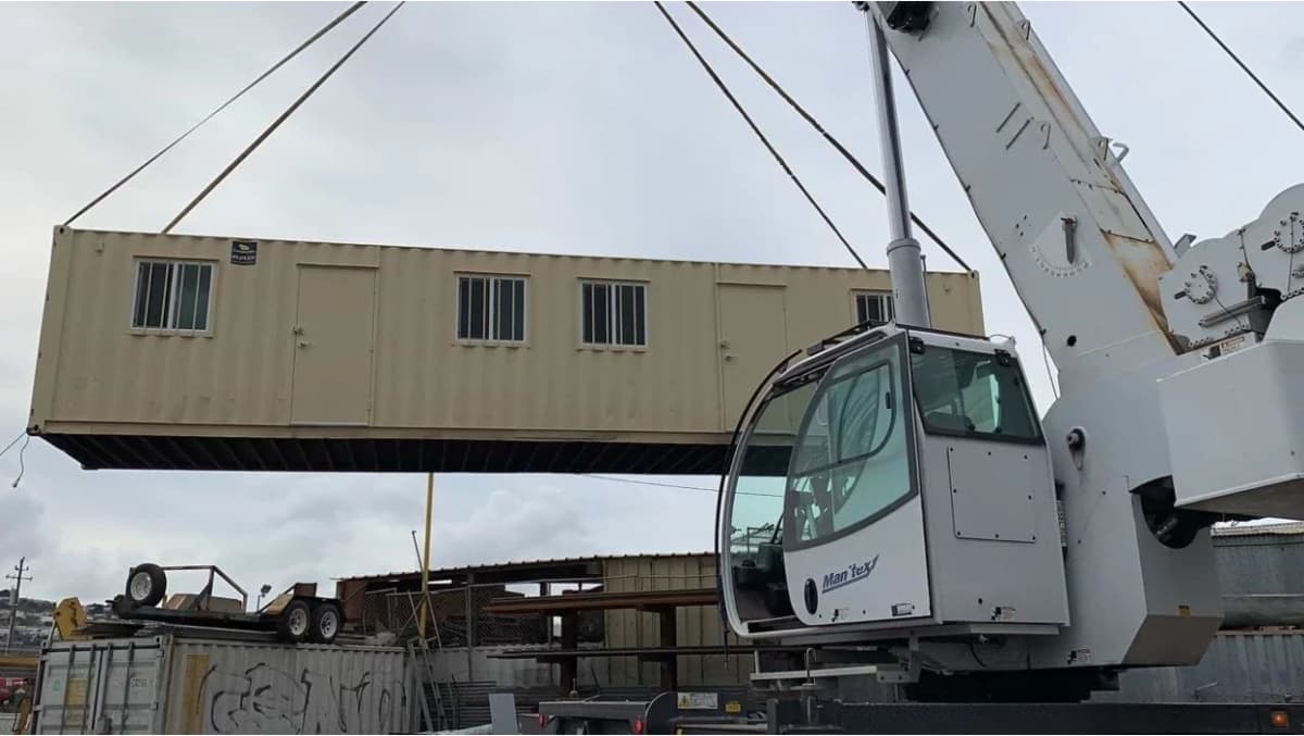 Large mobile crane lifting a beige shipping container with windows and doors.