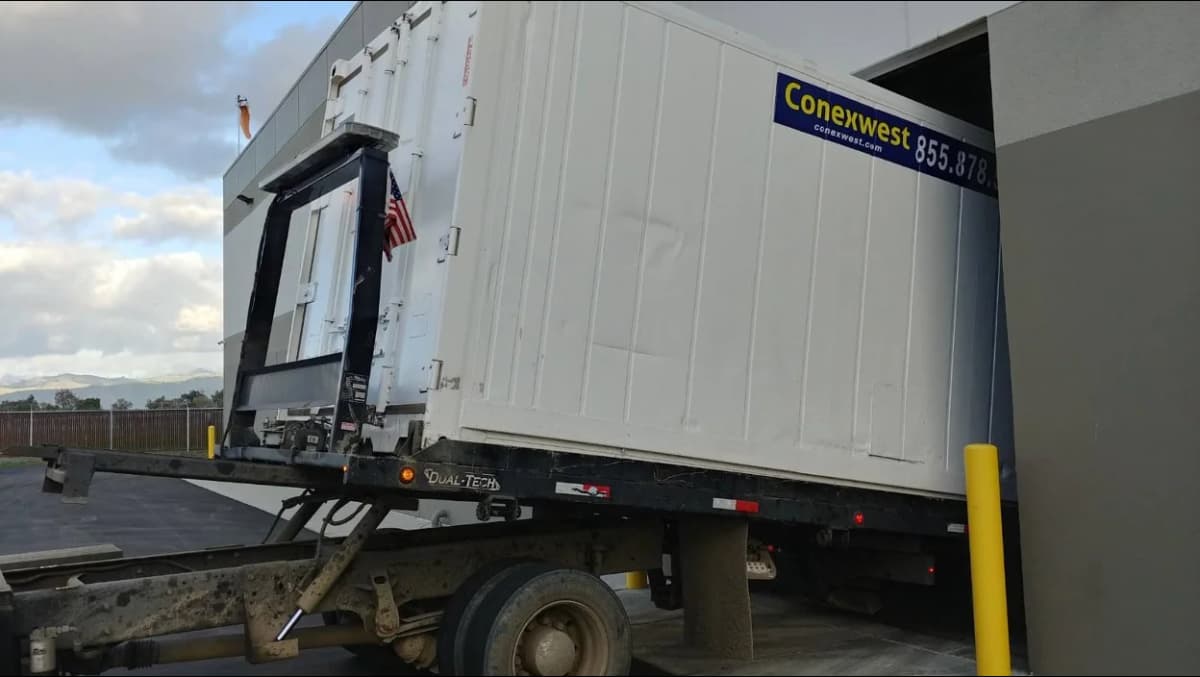White Conexwest shipping container being unloaded from a delivery truck.