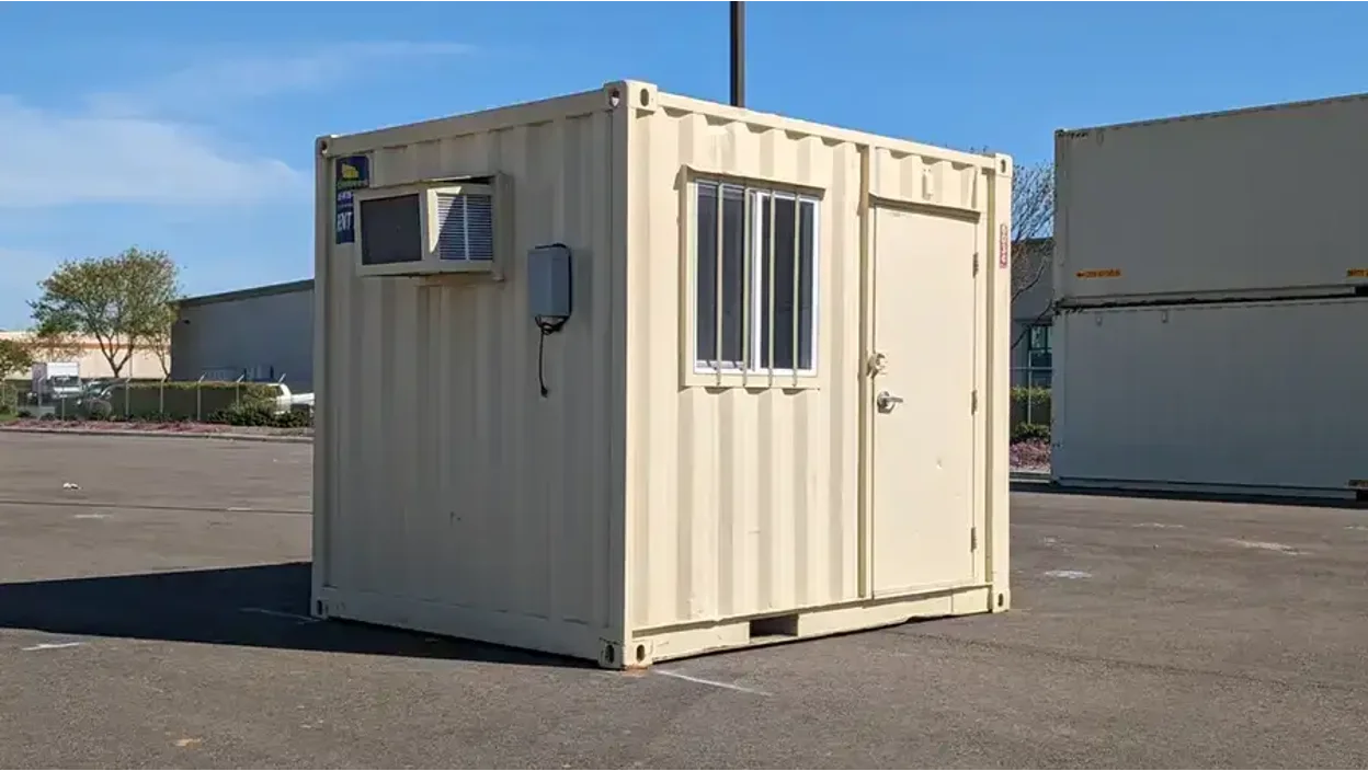 A beige portable Conexwest office container with a window, door, and wall-mounted air conditioning unit, placed in a parking lot.