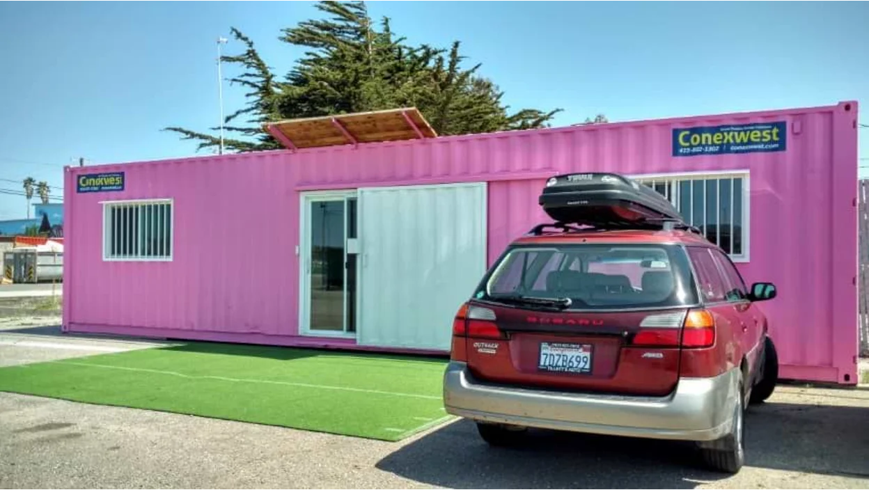 Bright pink shipping container converted into an office with sliding glass doors and windows, labeled “Conexwest,” with a red Subaru Outback parked in front on a patch of artificial grass.