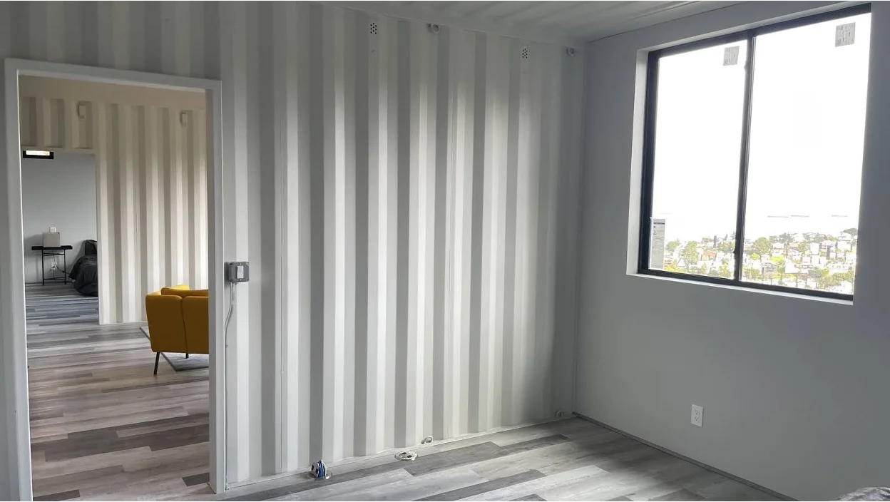 Interior of a modern container home with light gray corrugated metal walls, large window overlooking a coastal neighborhood, and wood-look flooring; an open doorway reveals a yellow chair and additional living space.