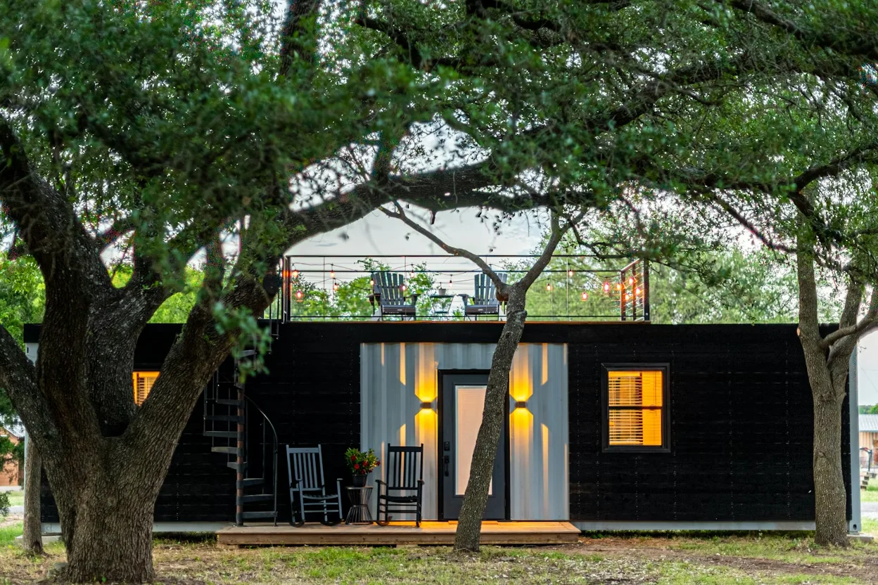 Modern black-and-gray shipping container home surrounded by large trees, featuring a small porch with rocking chairs, warm exterior lighting, and a rooftop deck with string lights and outdoor seating.