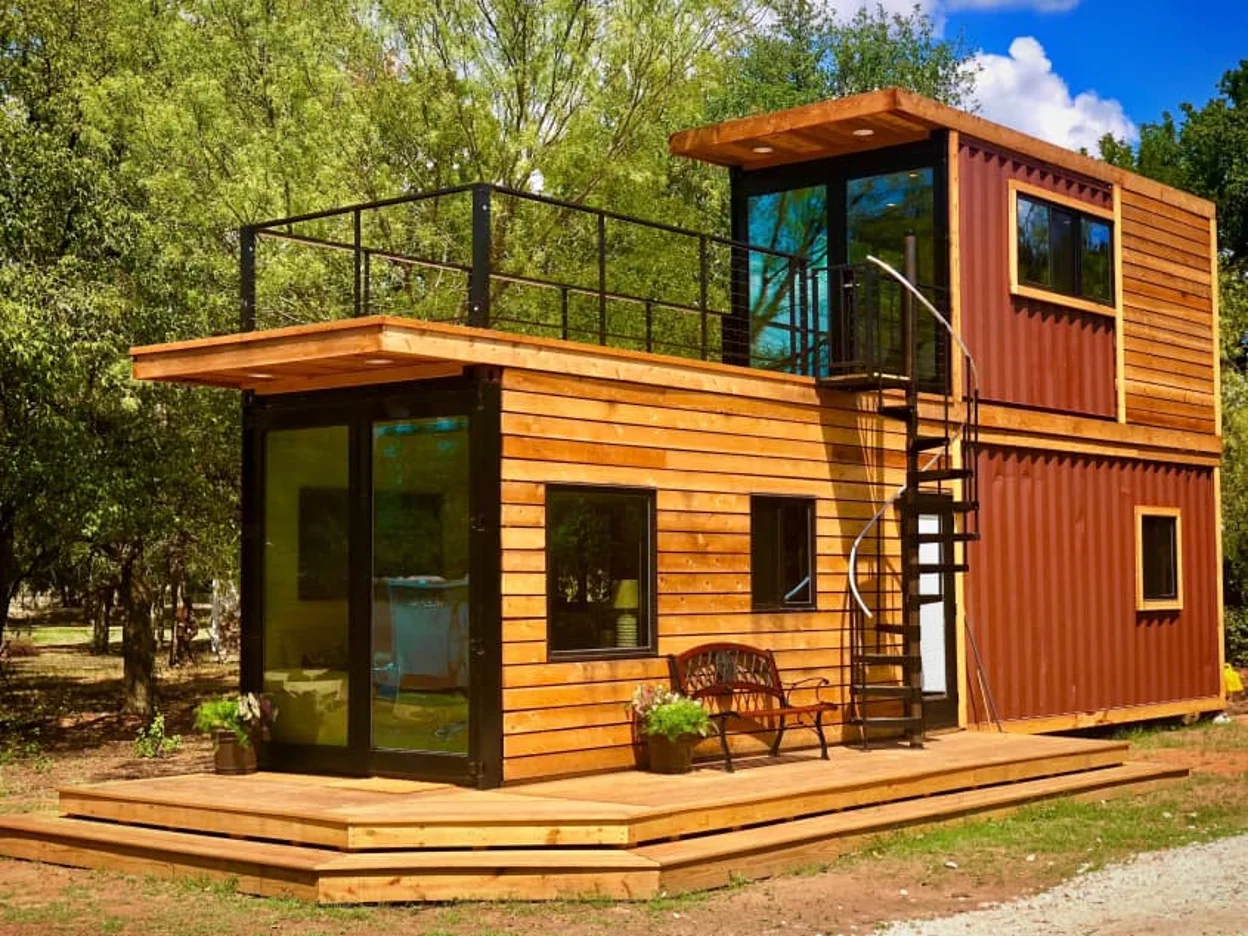 Modern two-story tiny home made from repurposed shipping containers, featuring wood siding, large windows, a spiral staircase, rooftop deck with railing, and a front porch.