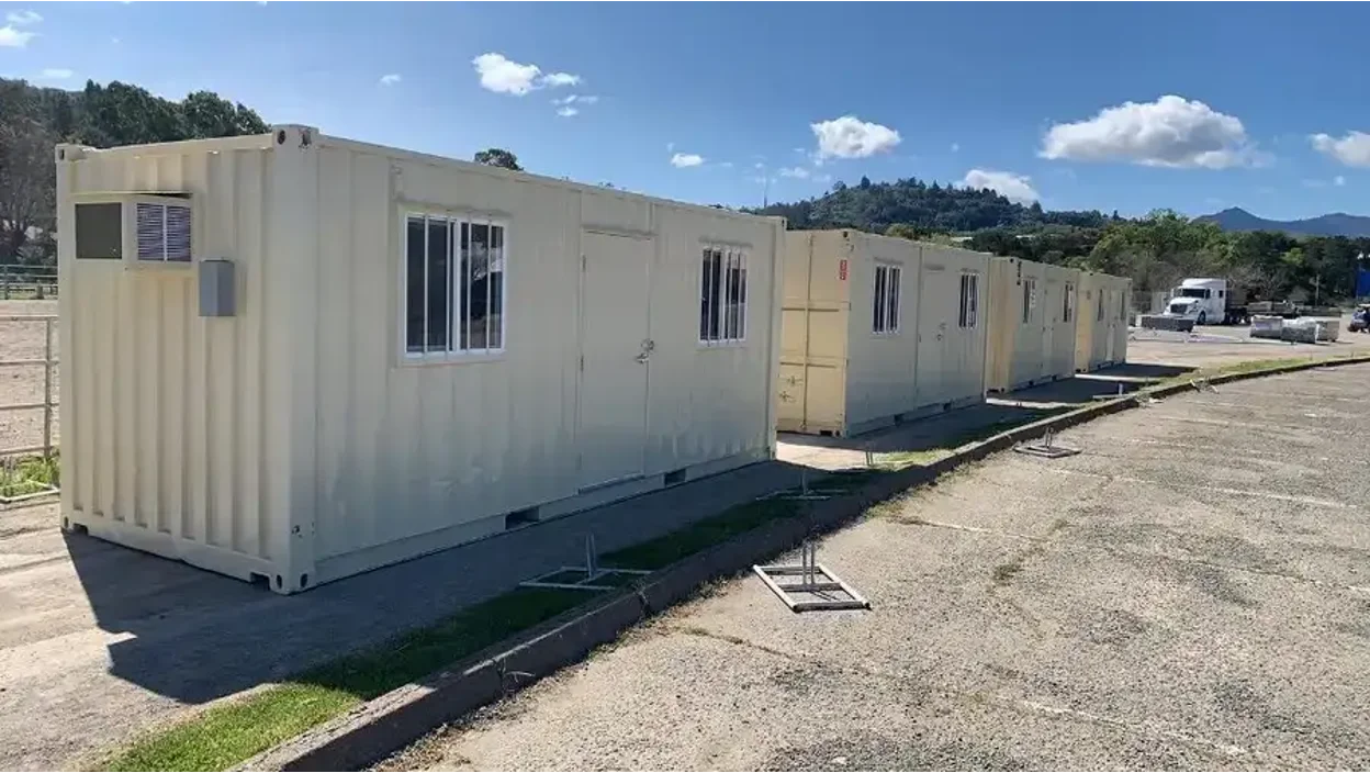 Row of beige portable container offices with windows and air conditioning units, lined up on a paved lot